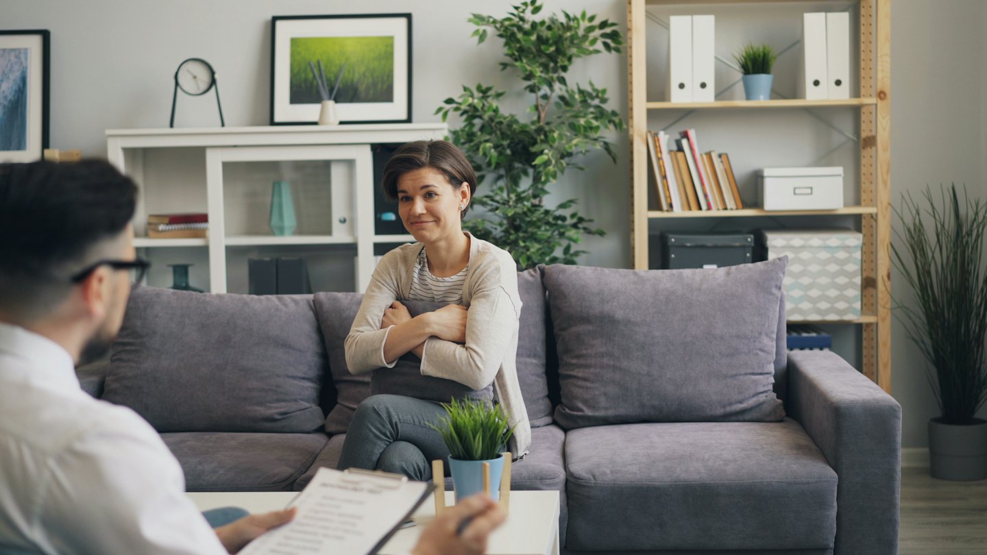 Therapist takes notes while client sits on sofa with cushion.